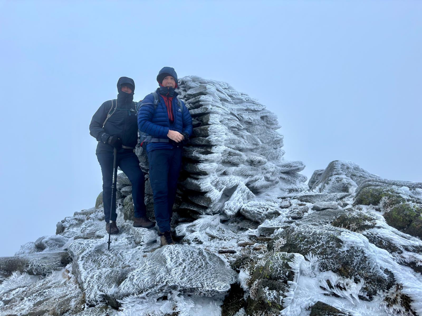 Two hikers in winter gear stand on a snowy, rocky mountain summit beside a frost-covered cairn. The scene is cold and adventurous.