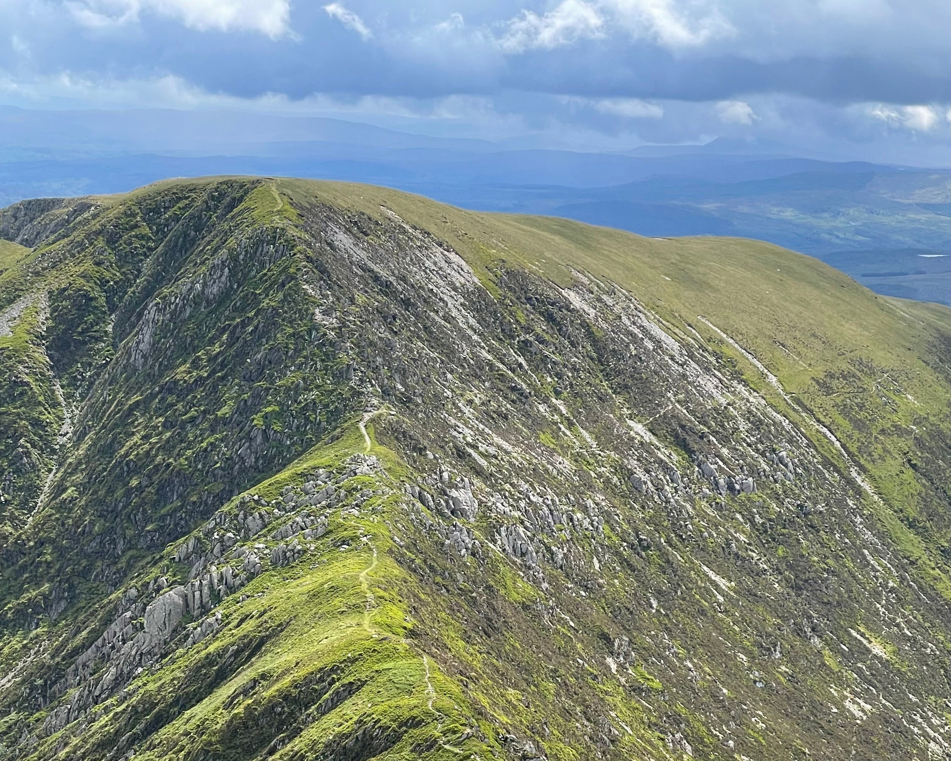 A rocky mountain ridge under a partly cloudy sky. The lush green slopes, dotted with rocks and a narrow trail, create a serene and majestic atmosphere.