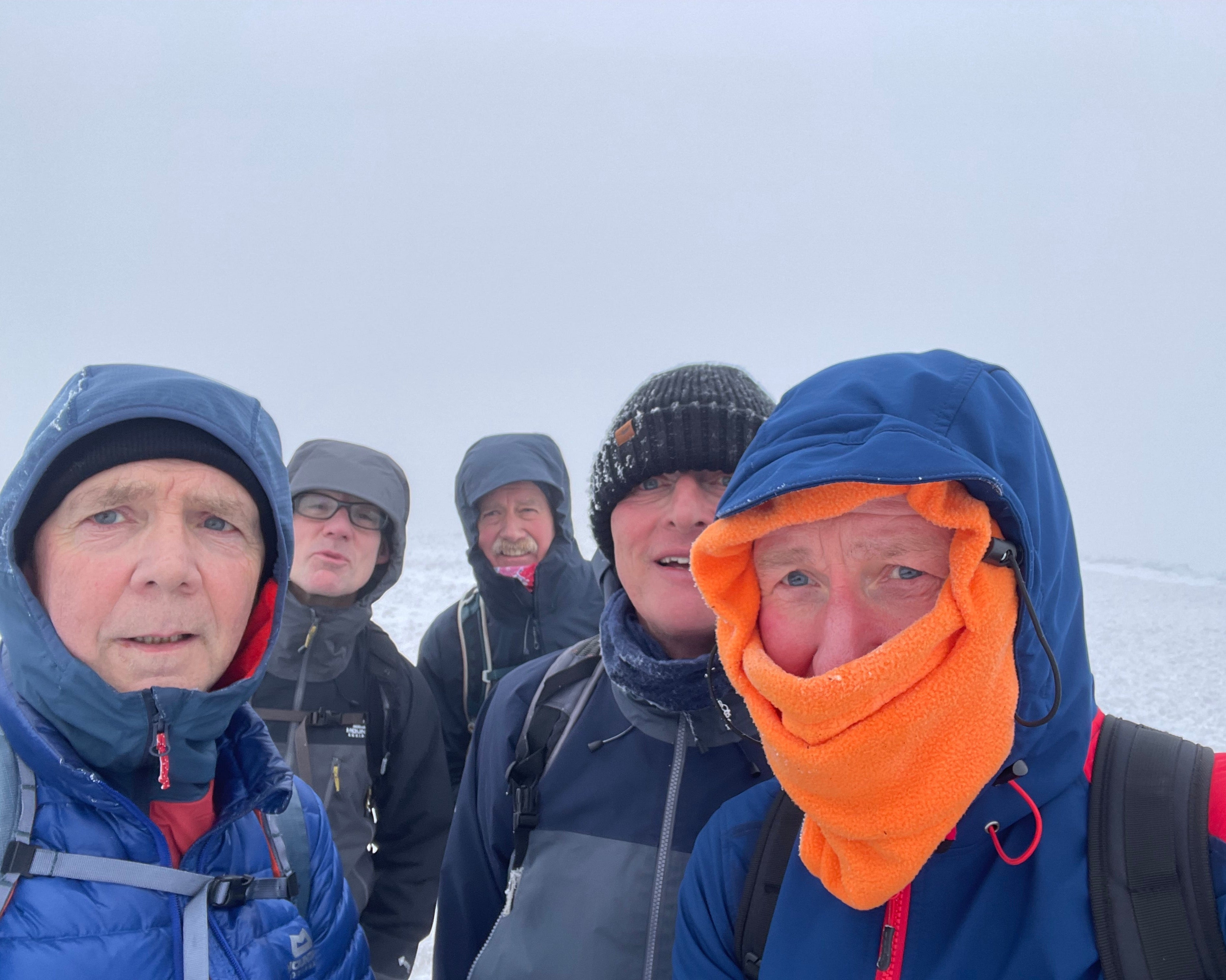 A group of five people bundled in winter gear, including beanies and scarves, stand closely together in a snowy, foggy landscape, looking content yet chilly.