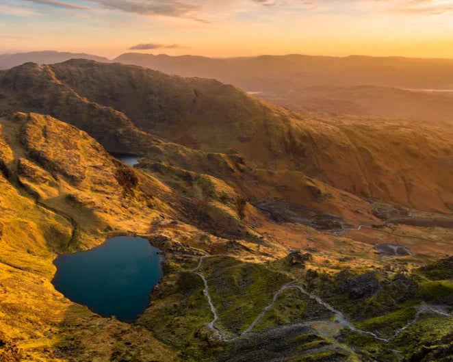 Sunrise over a mountainous landscape with a serene, blue lake nestled among rugged hills. The warm light casts a golden hue, creating a tranquil atmosphere.