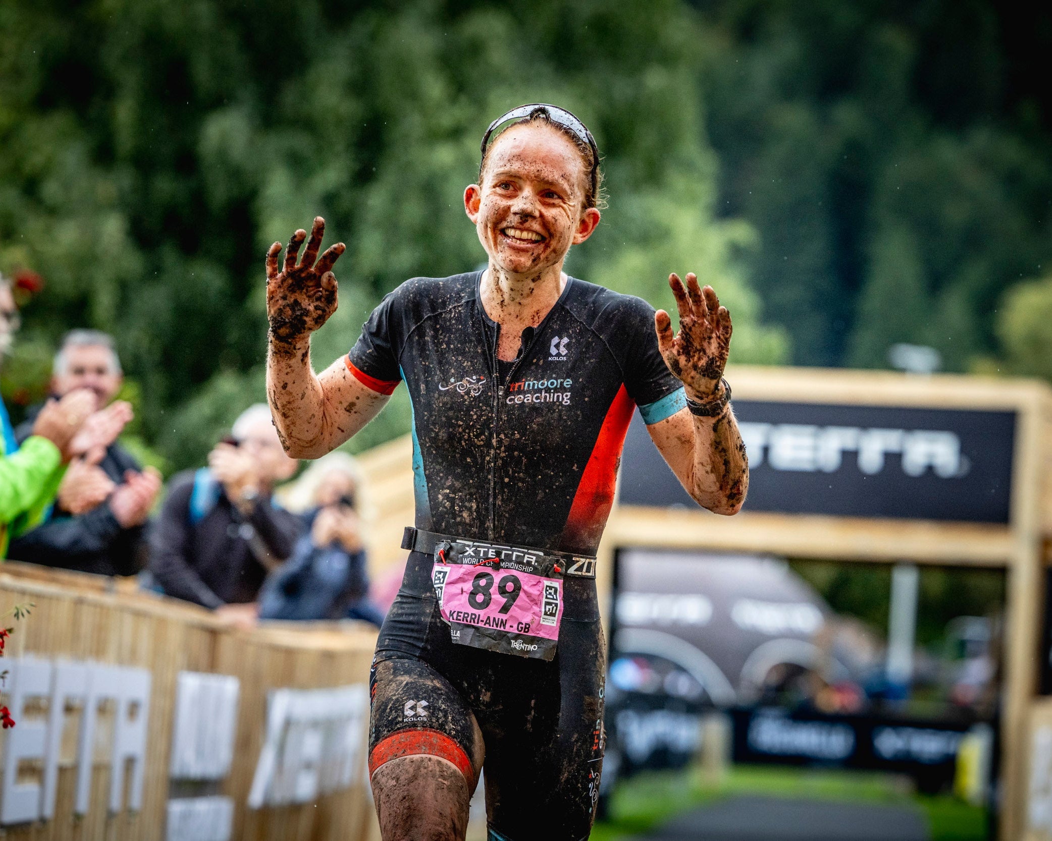 Athlete celebrating after a race with muddy hands and face, surrounded by spectators and event branding.