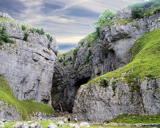Cave entrance between two rocky cliffs with greenery and a person in the distance.